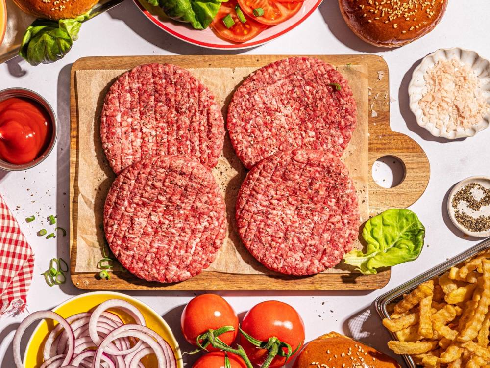 Fire rå hamburgerkjøttdeig på en skjærefjøl omgitt av grønnsaker og andre burgeringredienser