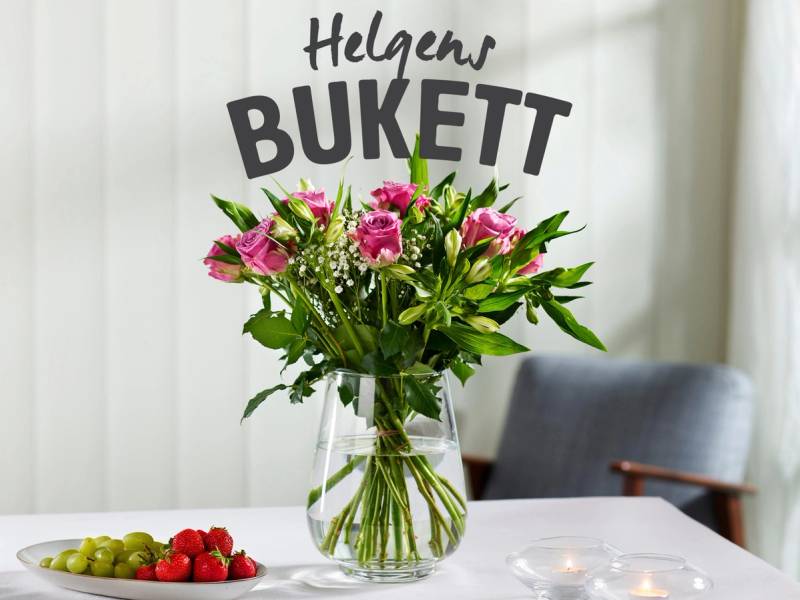 En bukett med rosa og hvite blomster i en glassvase på et bord, med en skilt som sier 'Helgens BUKETT' i bakgrunnen.