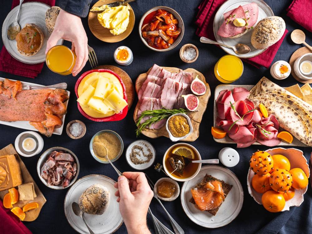 Et bord dekket med julefrokostmat, inkludert kjøtt, pålegg, egg, ost, frukt og drikke, sett fra oven.