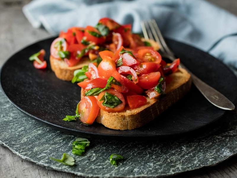 To skiver med bruschetta toppet med hakkede tomater, basilikum og olivenolje på en svart tallerken.