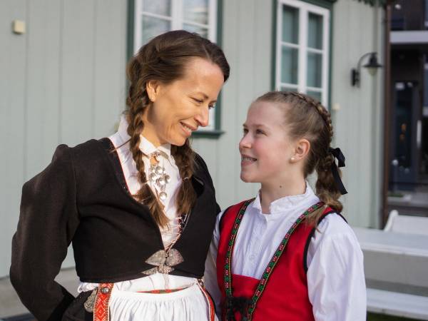 To jenter i tradisjonelle norske bunader med fletter, står sammen utenfor en bygning med store vinduer.