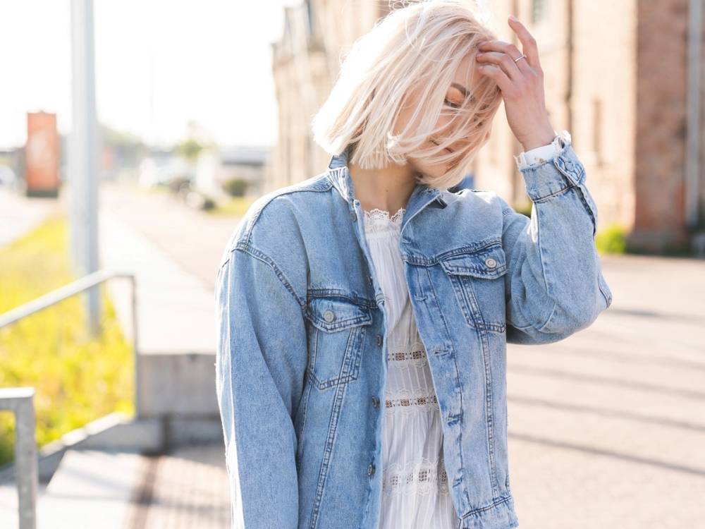 En ung kvinne med blondt hår som står utendørs og holder hånden i håret, iført denimjakke og lys skjorte.