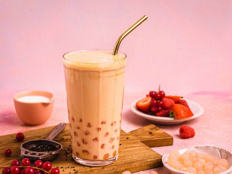 Et glass med melkete og et sugerør, omgitt av bær og en liten kopp, på et bord med en rosa bakgrunn.