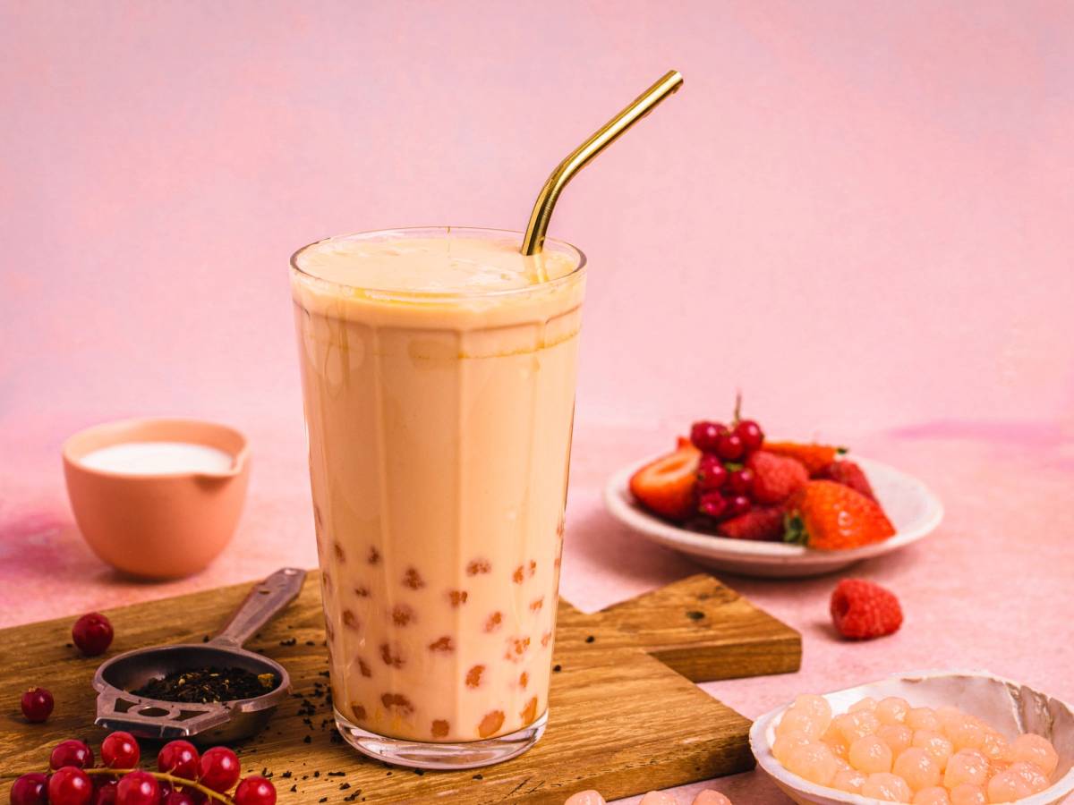 Et glass med melkete og et sugerør, omgitt av bær og en liten kopp, på et bord med en rosa bakgrunn.