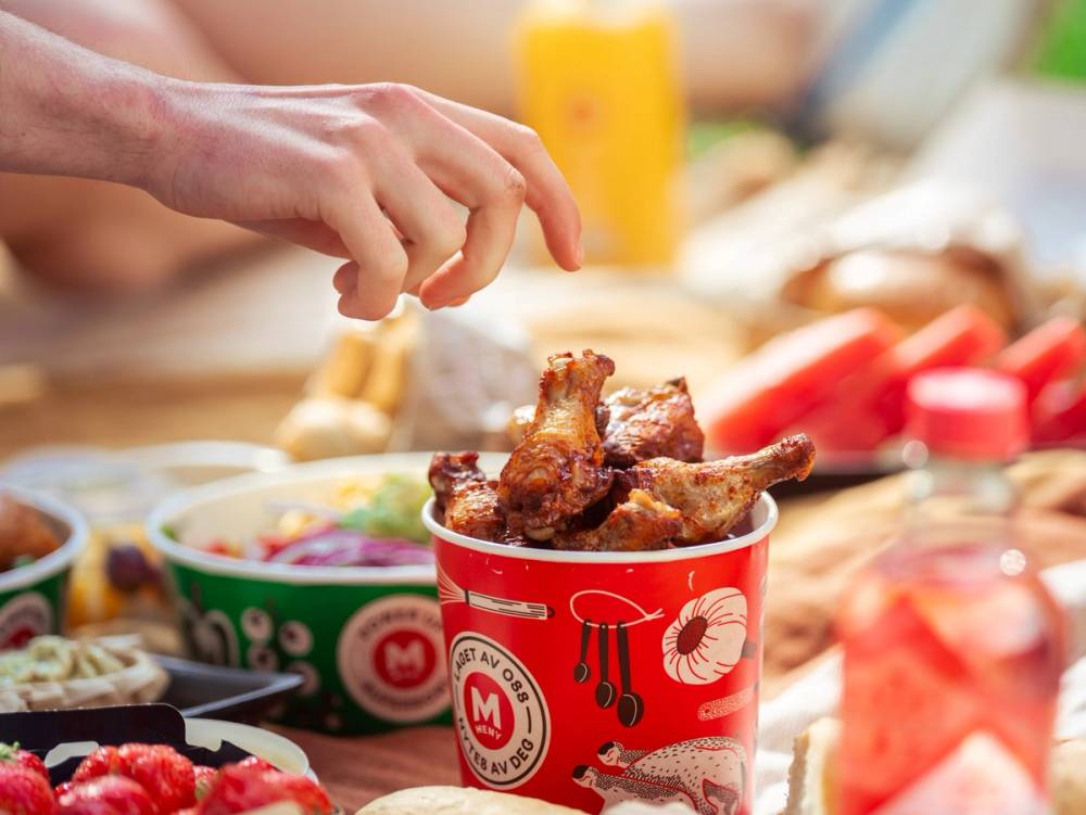 En person plukker opp en kyllingvinge fra en kopp med mat på et bord med ulike retter og ingredienser rundt.