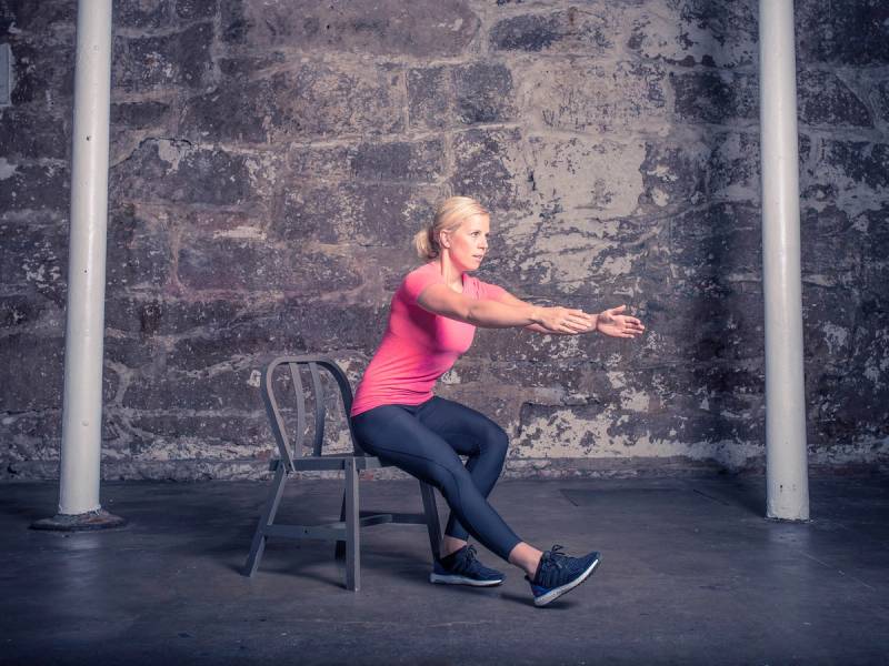 En person i rosa genser og blå bukser gjør en knebøy med et bein på en stol i et treningsrom.