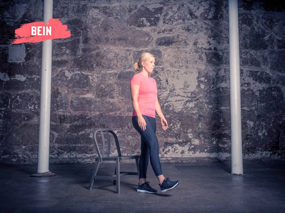 En kvinne utfører en knebøy med et bein oppstilt foran seg i et treningsrom med en murvegg i bakgrunnen.