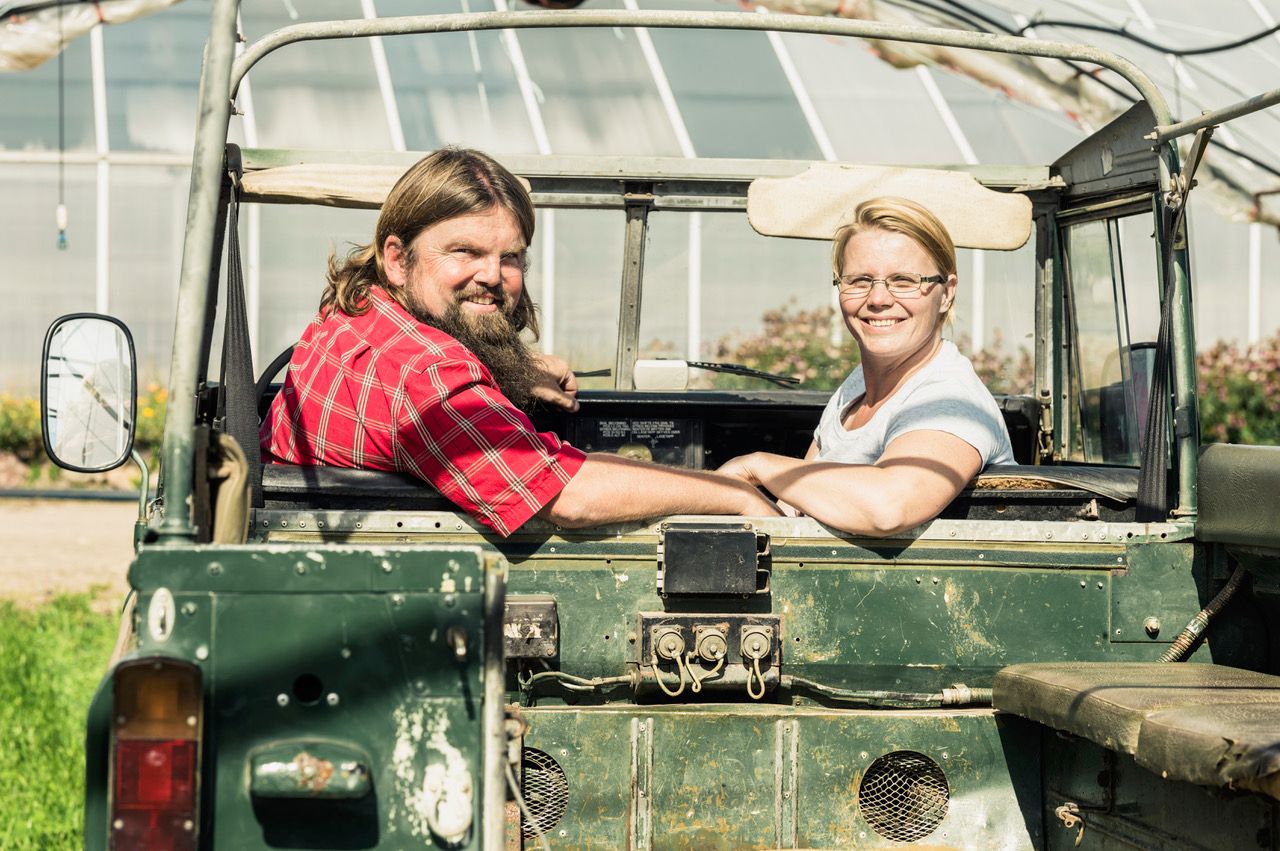 To personer sitter i en grønn åpen jeep, og snakker med hverandre utendørs på en solrik dag.