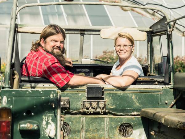 To personer sitter i en grønn åpen jeep, og snakker med hverandre utendørs på en solrik dag.