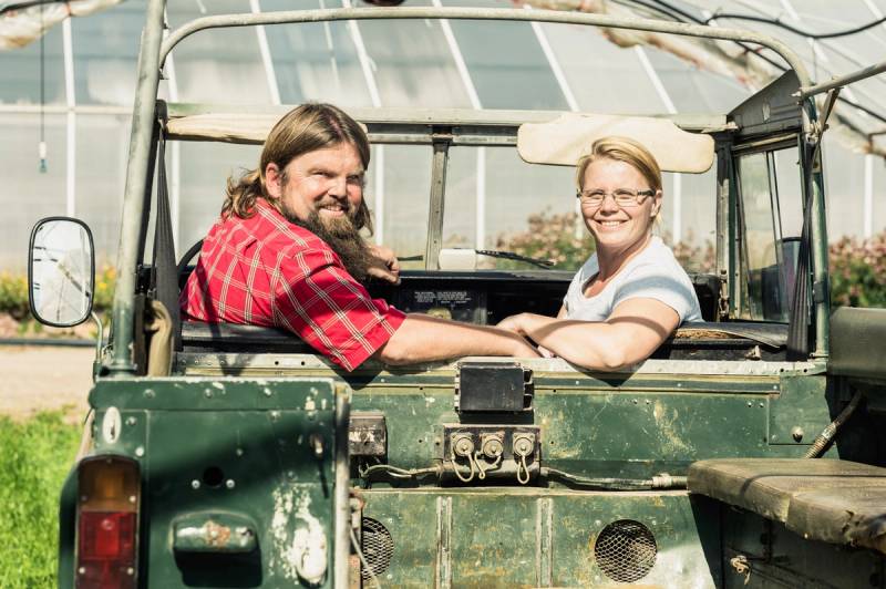 To personer sitter i en grønn åpen jeep, og snakker med hverandre utendørs på en solrik dag.