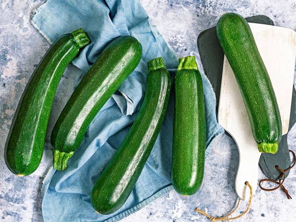 Fem zucchinis ligger på et bord med en kjøkkenkniv og en blå kjøkkenhåndkle, klar til å bli brukt i matlaging.
