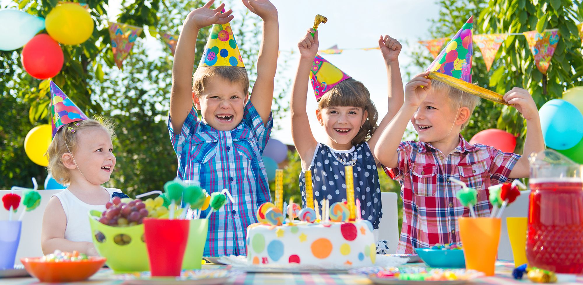 Barn feirer bursdag med kaker og pyntet bord utendørs, alle barna har på seg fargerike festhatter.