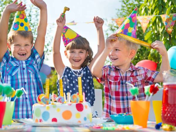 Barn feirer bursdag med kaker og pyntet bord utendørs, alle barna har på seg fargerike festhatter.