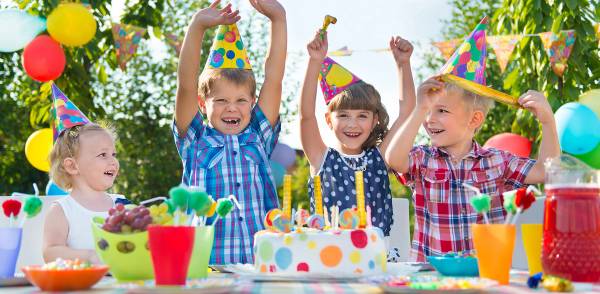 Barn feirer bursdag med kaker og pyntet bord utendørs, alle barna har på seg fargerike festhatter.