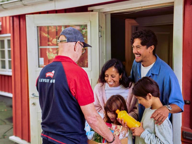 En mann i en rød og blå meny-levert-drakt deler ut mat til en gruppe barn utenfor en rød bygning.