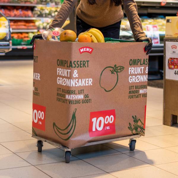 En person med svarte hansker som står bak en stor pappkasse med frukt og grønnsaker i en dagligvarebutikk, med prisskilt som viser 10 kroner.