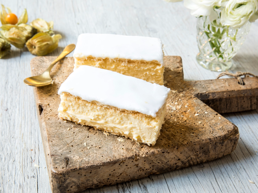 To napoleonskaker med hvit glasur på en treplate, omgitt av blomster og en liten skje.