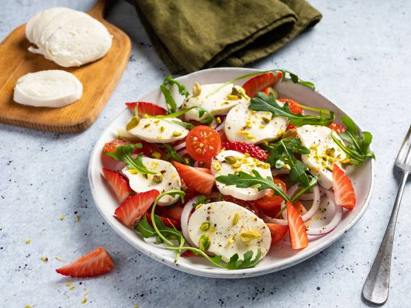 Mozzarella caprese salat med tomater, ruccola og basilikum på en hvit tallerken, med en skje og et grønt serviett ved siden av.
