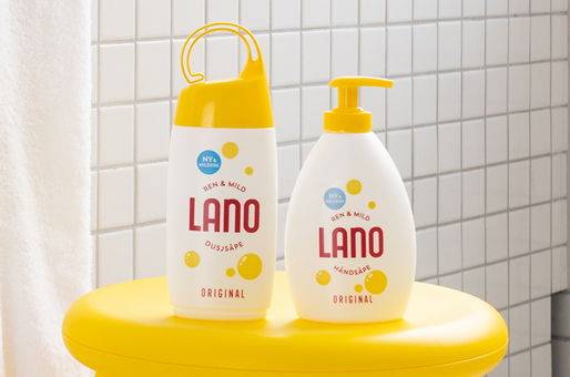 To Lano badeprodukter, en flaske med dusjsåpe og en med bodylotion, plassert på en gul overflate mot en hvit flislagt vegg.