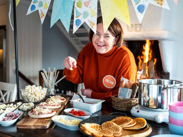Person som lager eller serverer mat ved et bord med ulike retter og snacks, med en peis i bakgrunnen.