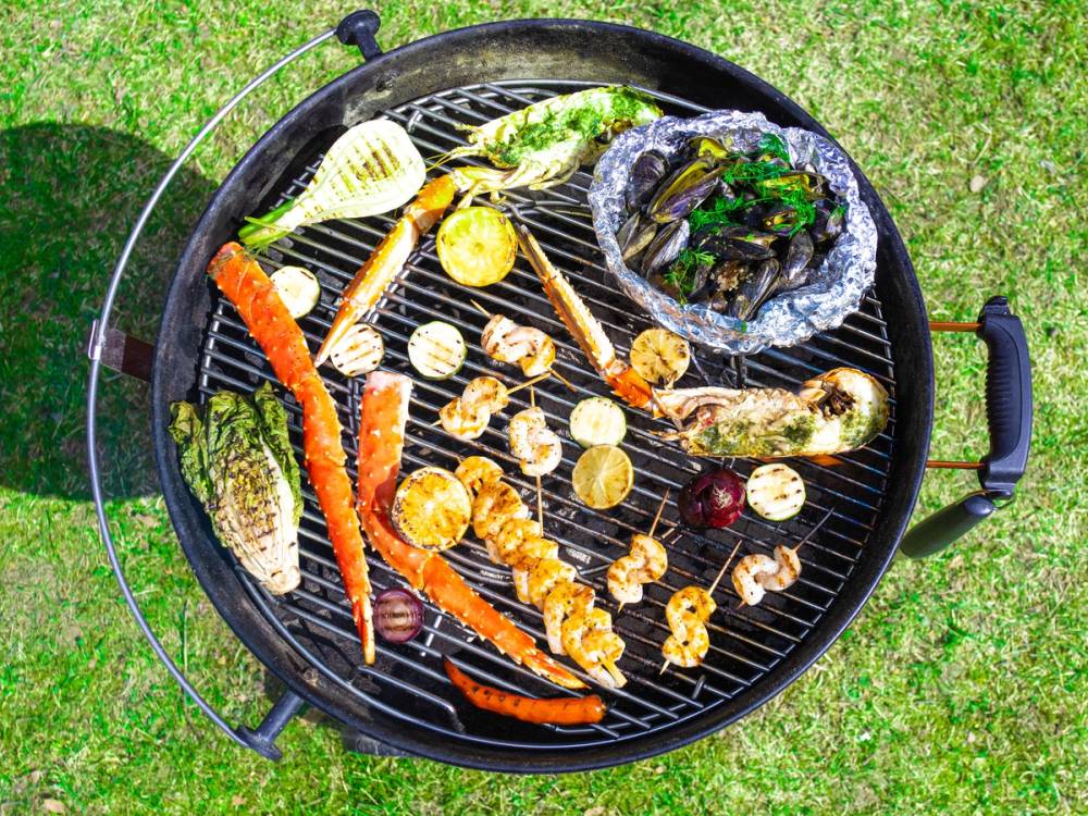 Grill med ulike fisker og skalldyr, inkludert reker, kamskjell og en fisk, på en gressplen.