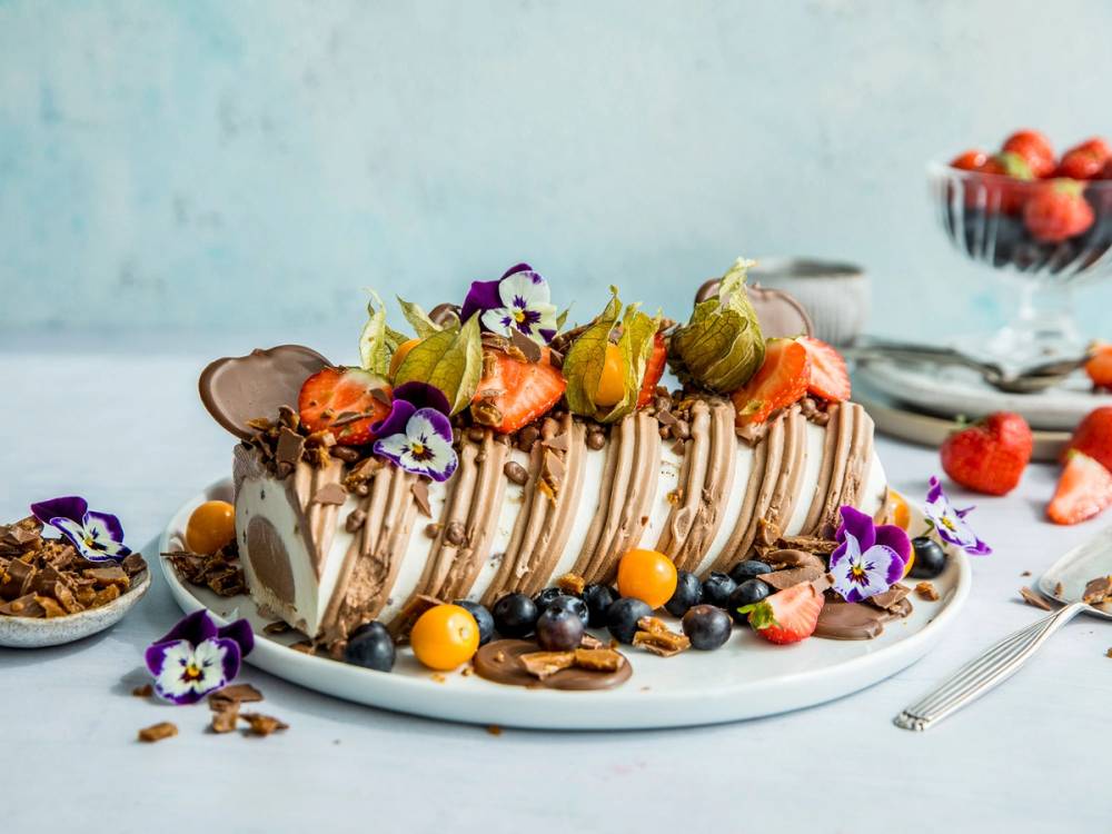 Daimiskake med sjokoladetrekk og pyntet med små blomster og frukt på et hvitt fat.