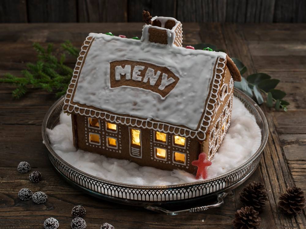 Et pepperkakehus med lysende vinduer og en liten pepperkakefigur foran på et fat med snøaktig pynt.