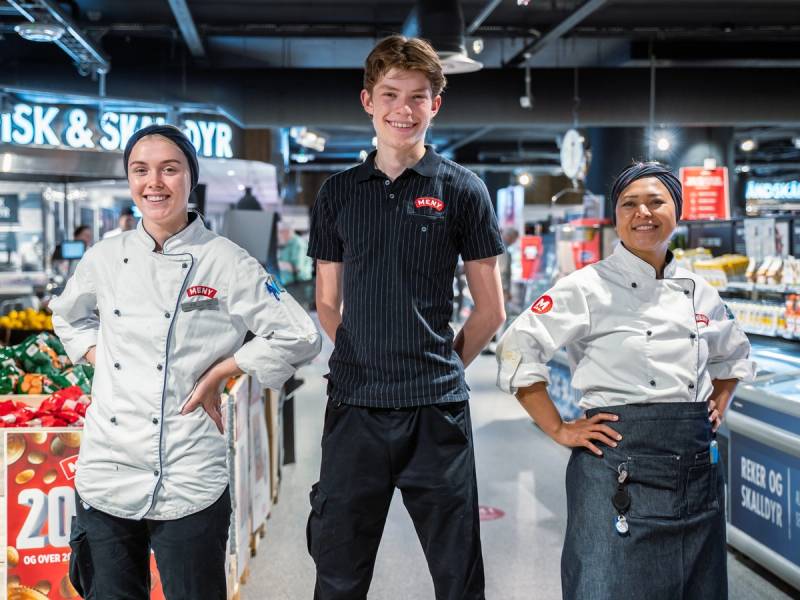 Tre ansatte i menyuniformer står i en dagligvarebutikk med frukt og grønnsaker i bakgrunnen.