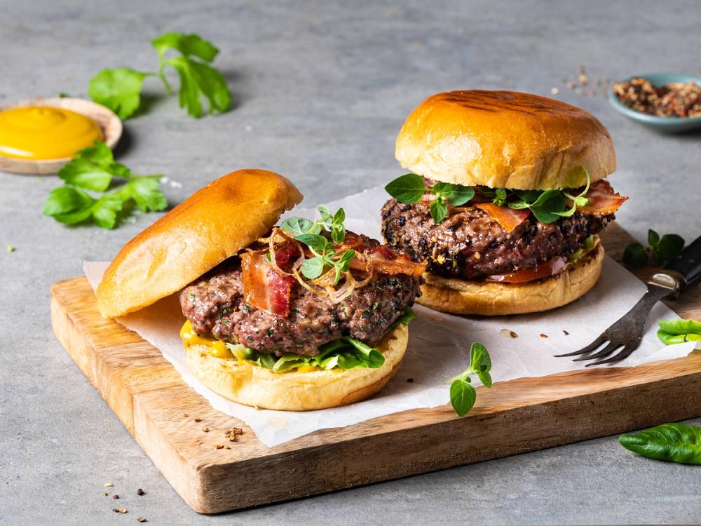 Hjemmelagde hamburgere med saftig kjøttdeig, salat og brød på en skjærebrett, omgitt av urter og krydder.