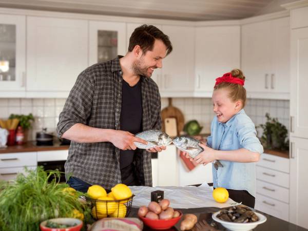En voksen og et barn deler en middag i kjøkkenet, med frukt og grønnsaker på benken foran dem.