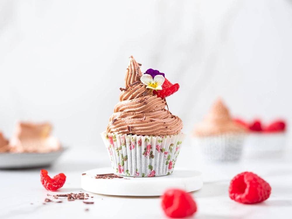 En cupcake med sjokoladefrosting og små spiselige blomster på toppen, omgitt av flere små kaker og bringebær på et hvitt bord.