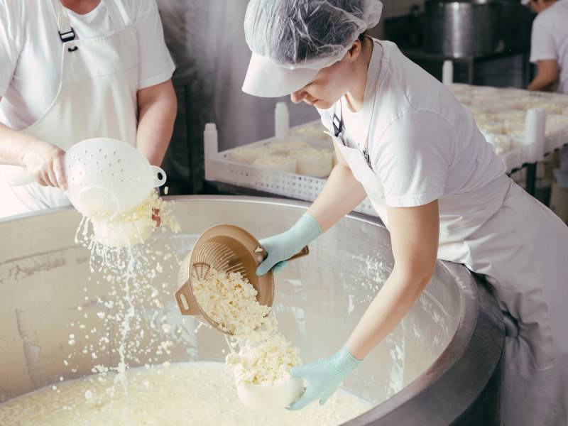 En person heller ris i en stor rund beholder, mens andre personer ser på, i et kjøkkenmiljø.