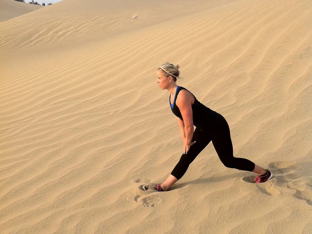 En kvinne i svart treningstøy som går opp en sanddyne i ørkenen, med sand som glir under føttene hennes.