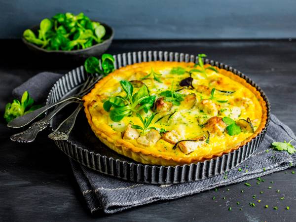 En gylden kyllingpai med grønne urter på toppen, ser ut til å være i en rund, svart paiform på et mørkt bord.