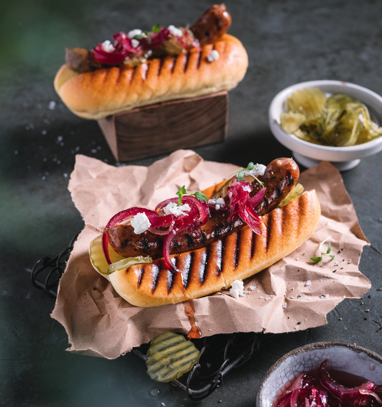 To pølser med rødløk, sennep og annet tilbehør i brød, servert på en tallerken med tilbehør i små skåler.