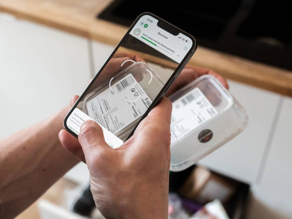 En person tar et bilde av en plastbeholder med mat på kjøkkenet med en smarttelefon.