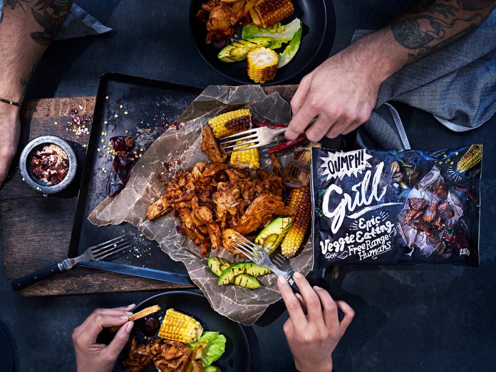 To personer som deler en tallerken med Oumph-mat, med mais og grønnsaker, på et bord med en pakke av Oumph-produkter i bakgrunnen.