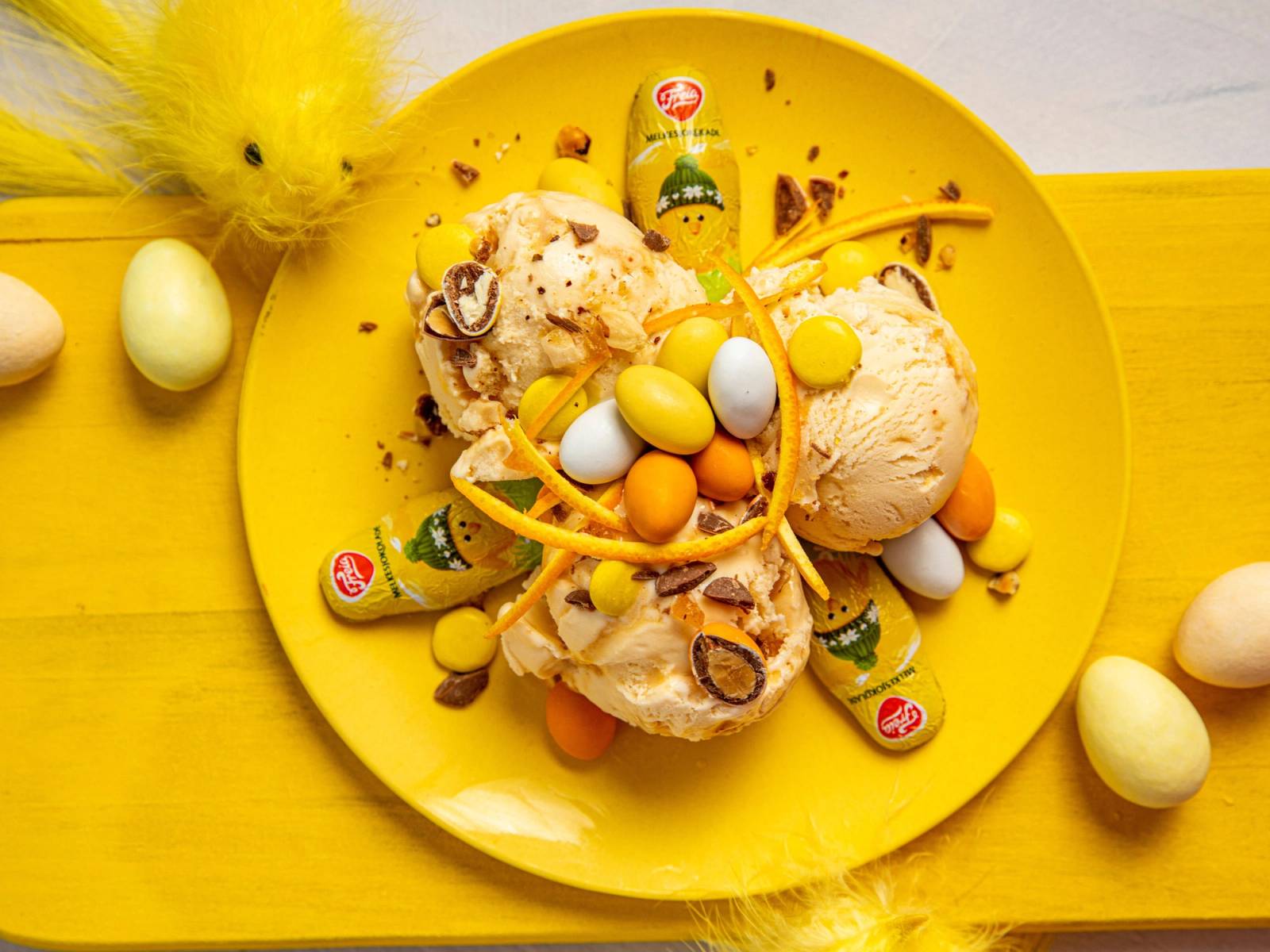 På en gul tallerken ligger iskrem med påskegodteri, sjokoladeegg og sitrusstrimler, omgitt av flere påskeegg på en gul bakgrunn.