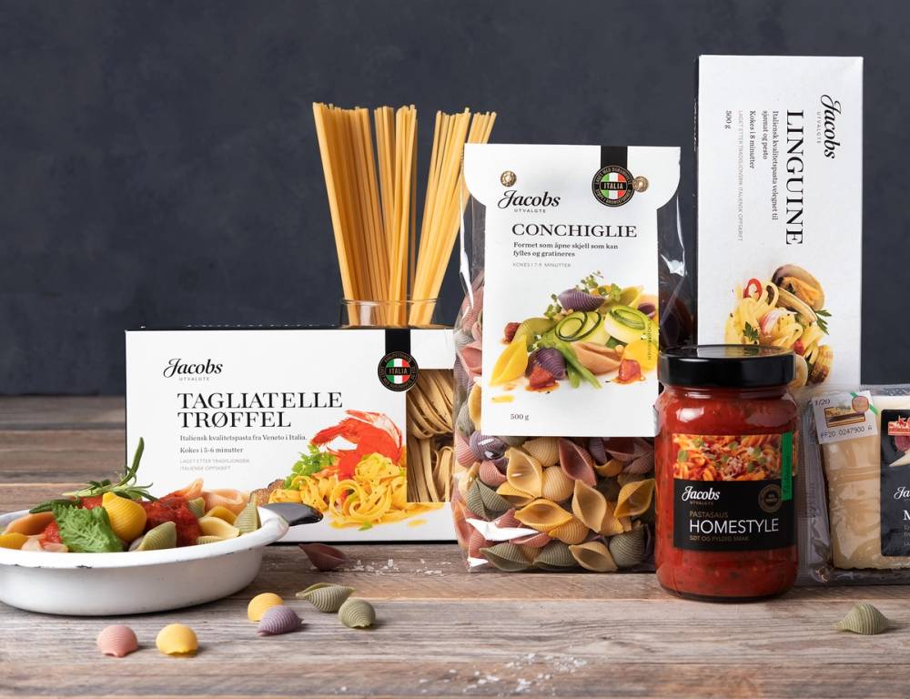 Ulike typer pasta og tilbehør fra Jacobs utvalgte pasta-serie, inkludert tagliatelle, conchiglie og andre varianter, med en bolle salat og en glass med tomatsaus.
