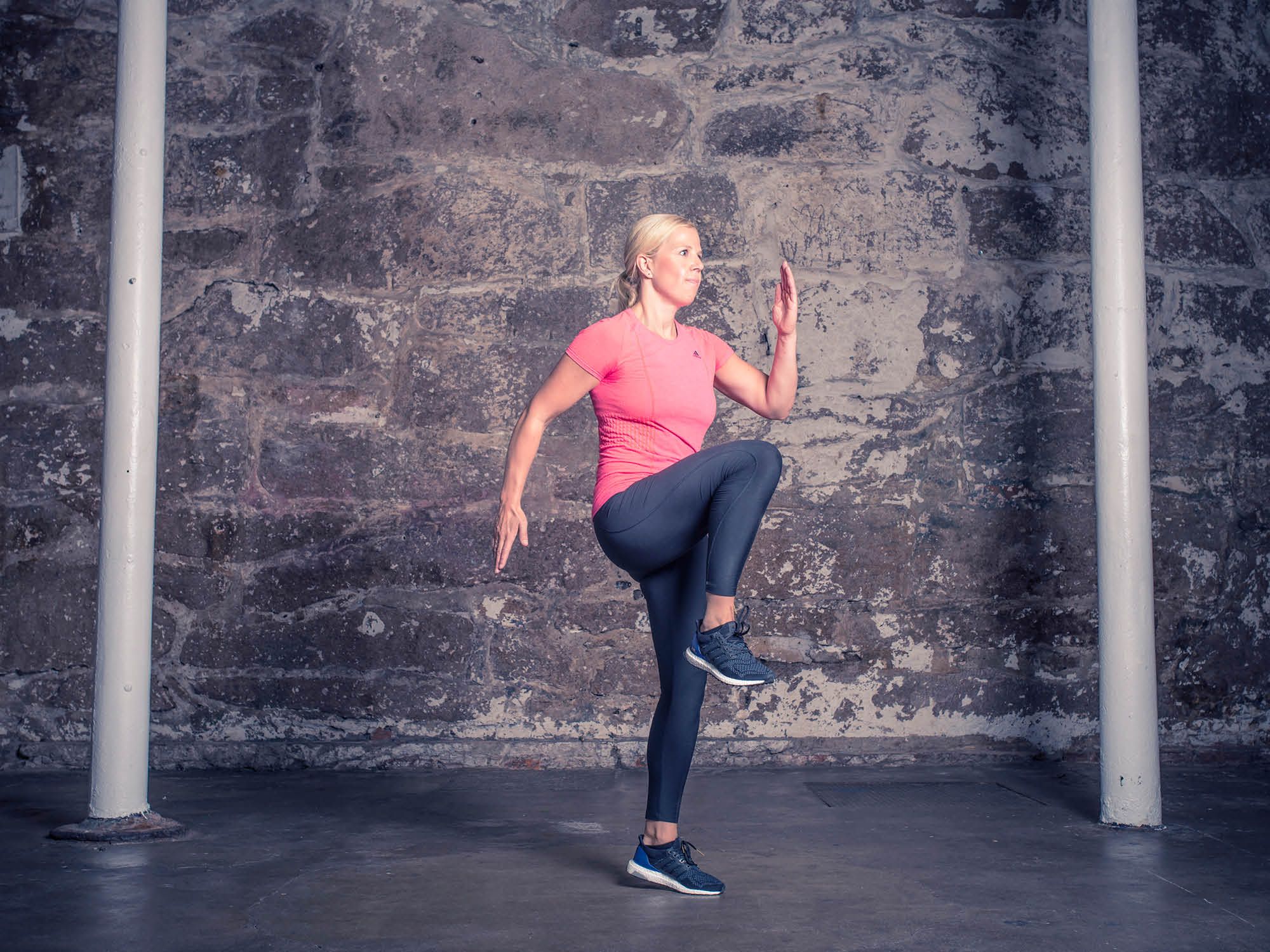 En person utfører høye kneløftøvelser i et treningsrom med en murvegg i bakgrunnen.