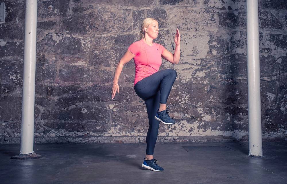 En person utfører høye kneløftøvelser i et treningsrom med en murvegg i bakgrunnen.