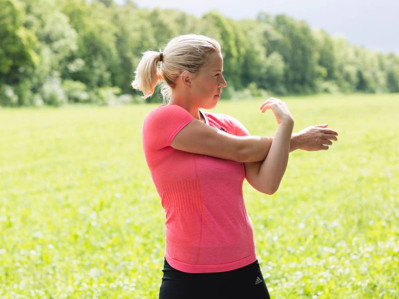 En kvinne i rosa treningstøy strekker armene utendørs i en grønn eng med trær i bakgrunnen.