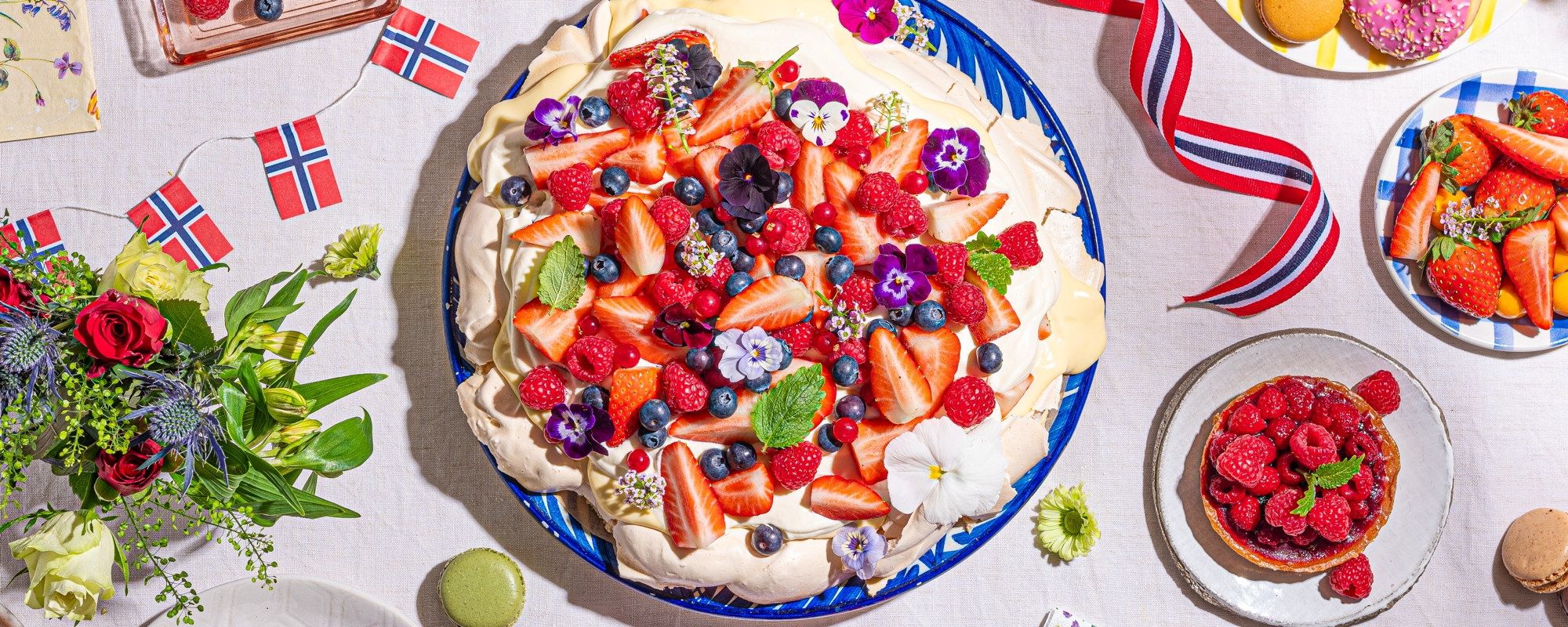 Et bord dekket med ulike desserter, inkludert en frukttårn med jordbær, blåbær og kiwi, samt små kaker og fruktfat, pyntet med norske flagg og sommerlige dekorasjoner.