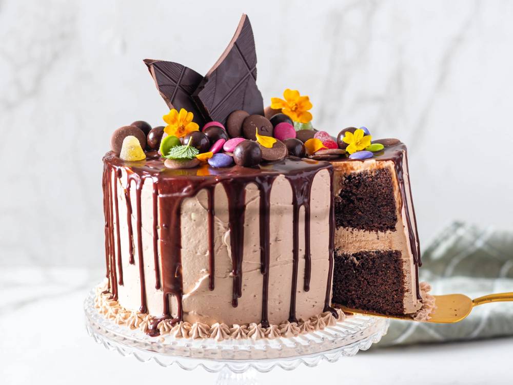 En sjokoladekake med drypp av sjokolade på kanten og pyntet med fargerike sjokoladekuler, små gule blomster og et stykke av kaken er skåret ut og ligger på en tallerken med en skje.