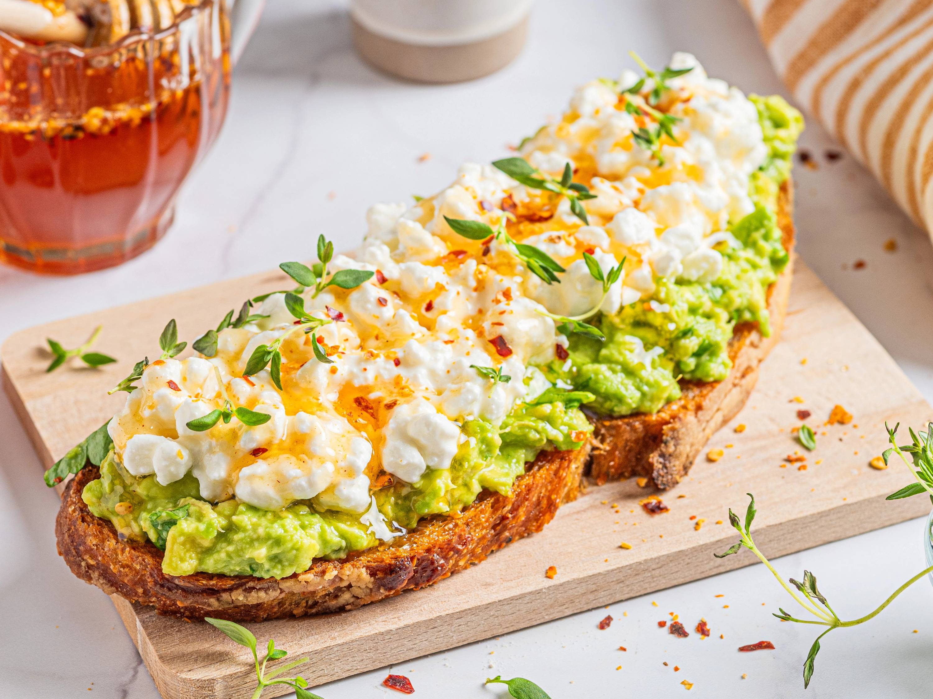 To rista brødskiver med avokadomos, cottage cheese og hot honey, pyntet med urter på en skjærebrett.