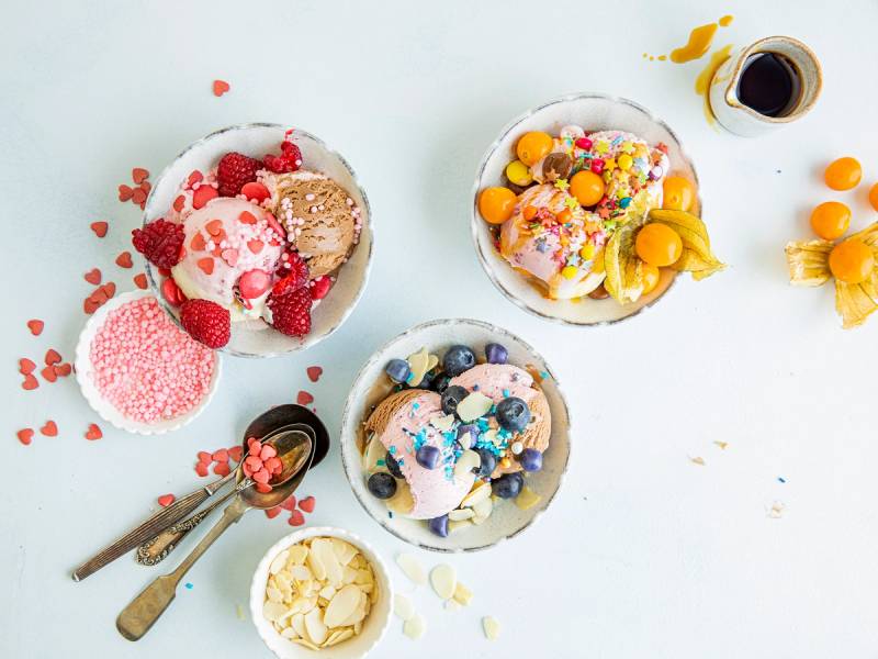 Ulike typer iskrem i skåler med bær, sjokolade og godteri, samt en kopp med kaffe på et hvitt bord.