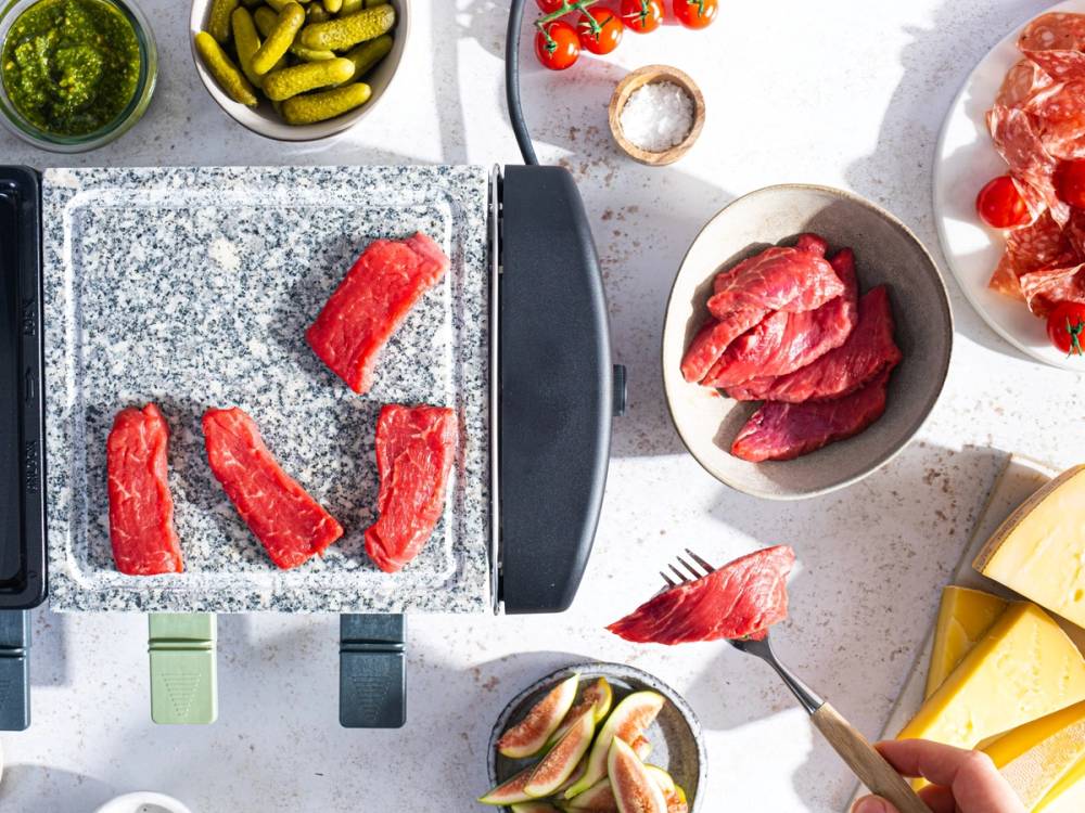 Raclette-plate med skiver av smeltet ost og ulike typer kjøtt, omgitt av tilbehør som pickles og grønnsaker