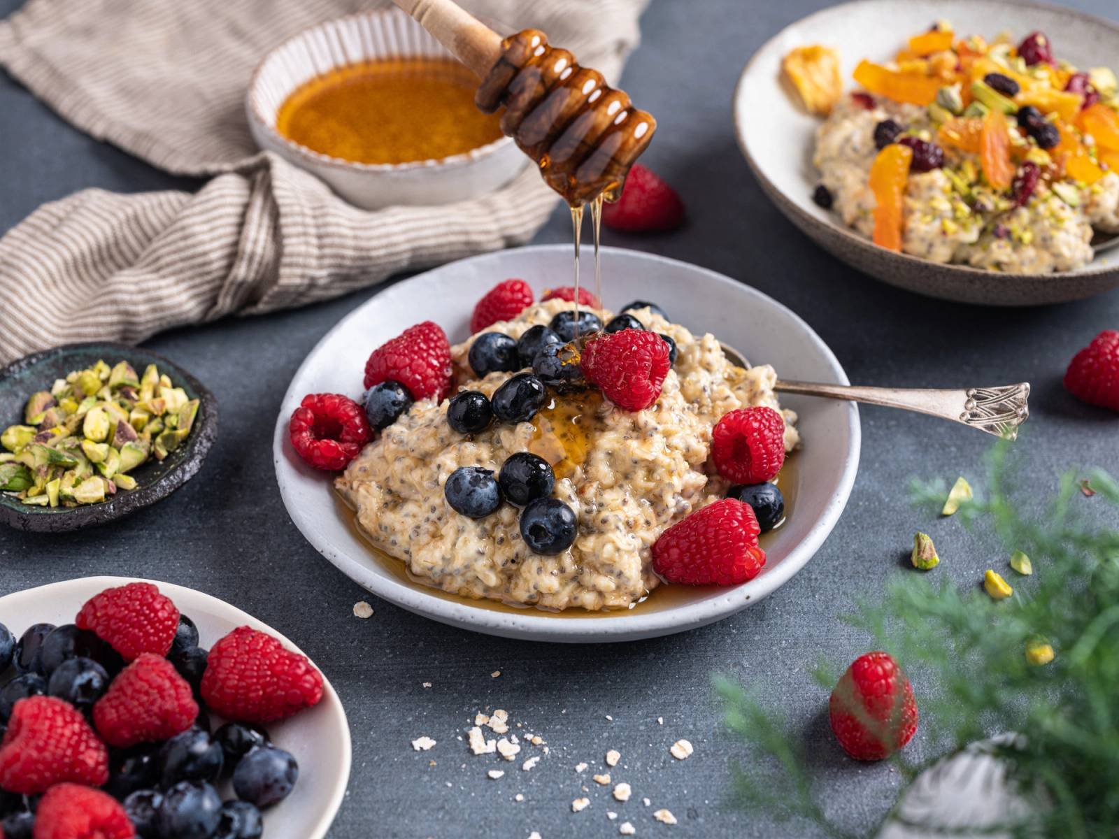Havregrøt med bær og honning på et svart fat, omgitt av flere små skåler med frukt og nøtter.