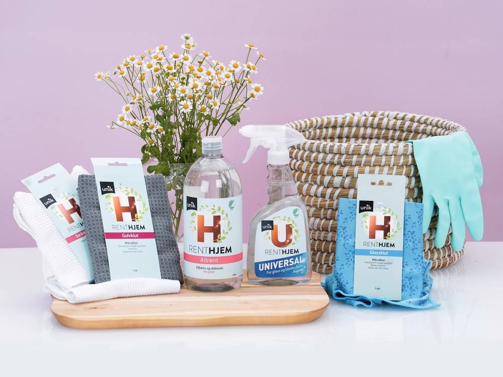 En samling rengjøringsprodukter, en kurv og vaskeklut på et bord med en vase med hvite og gule blomster i bakgrunnen.