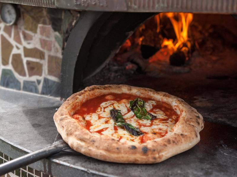 En pizza ligger på en pizzaspade foran en varm pizzaovn med åpen ild.